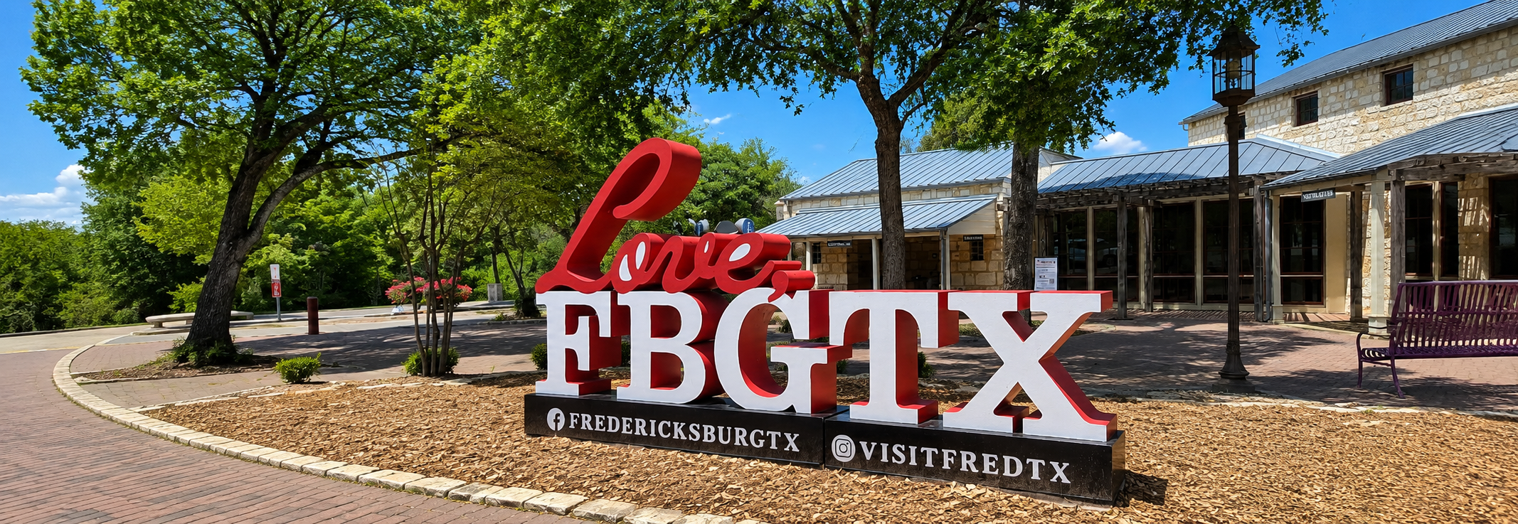 Large red and white 'Love FBGTX' sign in front of a limestone building with a metal roof and shade trees, with Fredericksburg tourism social handles along its base